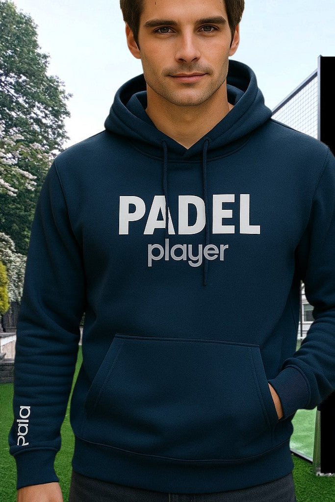 Man wearing a navy blue hoodie with the words 'PADEL player' printed on the chest in white. The sleeve features the brand name 'pala.' printed vertically. He is standing on a padel court with a net and trees in the background.