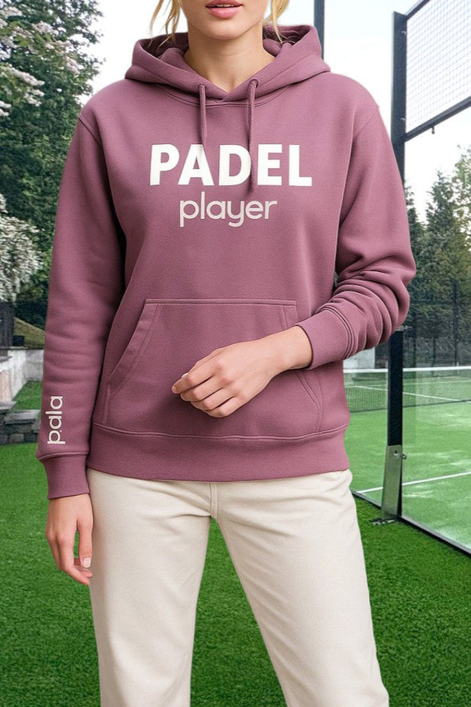 Woman wearing a dusty rose hoodie with the words 'PADEL player' printed in white on the chest and 'pala.' vertically on the sleeve. The hoodie has a front kangaroo pocket and drawstrings. She is standing on a padel court with green turf and glass walls