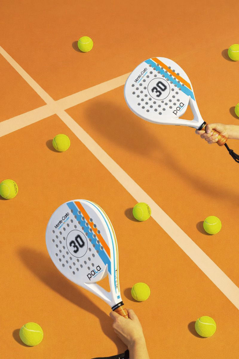 Two paddle tennis rackets with '30' branding held above a clay court with scattered tennis balls.